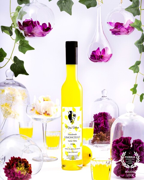 Limoncello bottle photographed in a "clear" scene with different shapes and sizes of glasses, with the yellow limoncello liquid in some of the glasses in contrast with the purple flowers in the scene.