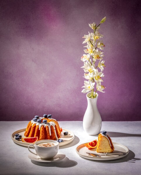 Perfect a sweet breakfast. Round shaped cake with icing and blueberries served on a plate with a slice of blood orange together with a cup of cappuccino