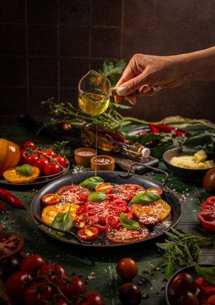Hand pouring golden olive oil from a glass measuring cup over sliced heirloom tomatoes with fresh basil and grated parmesan for a rustic, fresh salad or appetizer