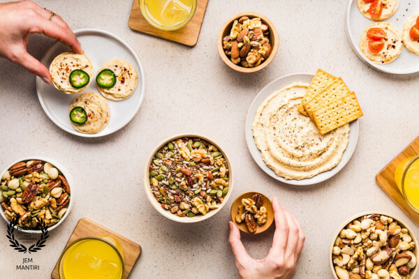 Flatlay photo of hummus with crackers, nuts, seeds, and refreshing drinks, taken for a food company...