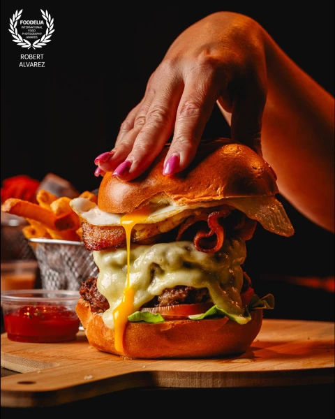 A hand presses down on a towering burger just before the first bite — and the egg yolk breaks on con...