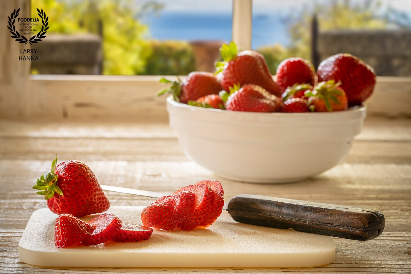 Cruising through the grocery store, the beauty of the fresh strawberries caught my eye.  I had just made a new white wood surface for my food shots and immediately though of creating a fresh morning look of strawberries being cut by the kitchen window.  I made both the surface and the window frame and layered in a view I had captured on a recent trip to Italy.