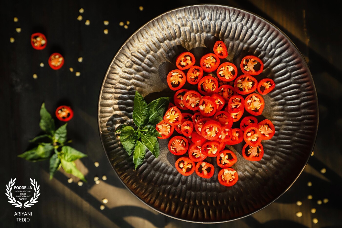 Dare to be hot and beautiful! Slices of fresh red chili peppers garnished with fresh chili pepper leaves from the garden. A gobo is used to give an ornamented shadow to the rustic wooden table underneath the raised plate.