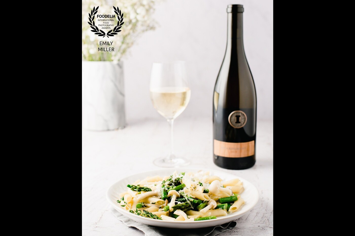 White plate with Vegan Asparagus Mushroom Pasta with Vegan Parmesan Cheese accompanied by a glass of Chardonnay and wine bottle in a bright and airy style with white background and green and white flowers in a marble vase.