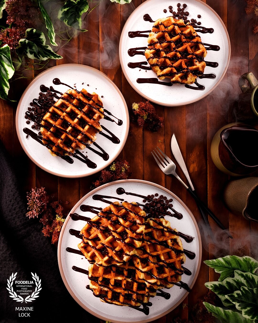 Plates of chocolate waffles photographed on a wooden table, using crockery as decorations but also using green leaves and smoke to create more of a scene.