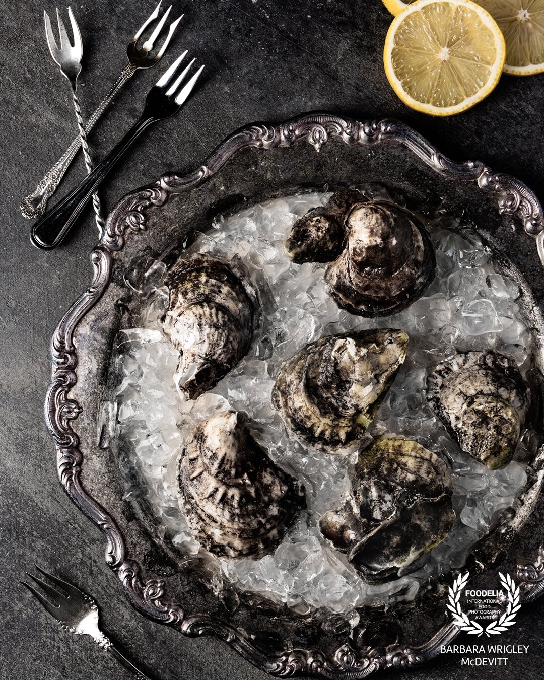 Oysters are one of my favorite subjects to photograph - with all their texture they are so photogenic.  I wanted to create a monochromatic color scheme and displayed these oysters in an antique silver platter that complemented the grey color of the oysters.  I chose a dark grey background and used antique seafood forks to carry through on the color scheme.  A touch of color was added by the lemons to break up the monochromatic color scheme.