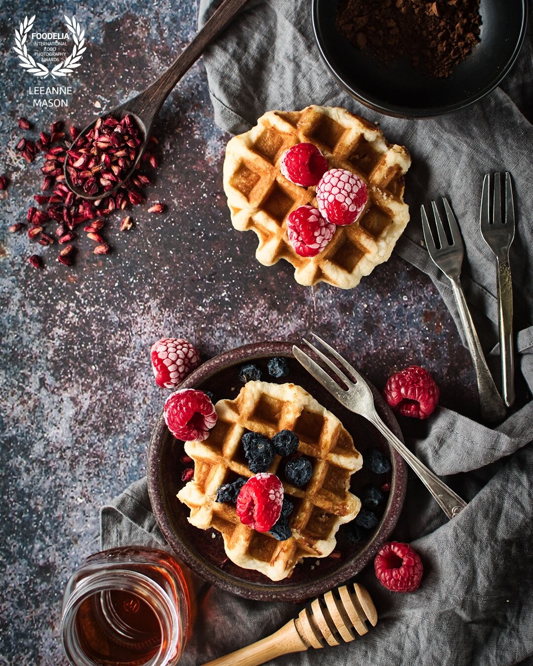 Delicious waffles with pomegranate seeds, berries, and maple syrup and a little bit of chocolate on the side the only decision left is whether to serve them with ice cream or yoghurt.  The lines in the image leads the viewer around the frame taking in all the details, from the texture of the frozen berries to multiple forks indicating that this image is part of a larger scene, perhaps a table with more people and more waffles!