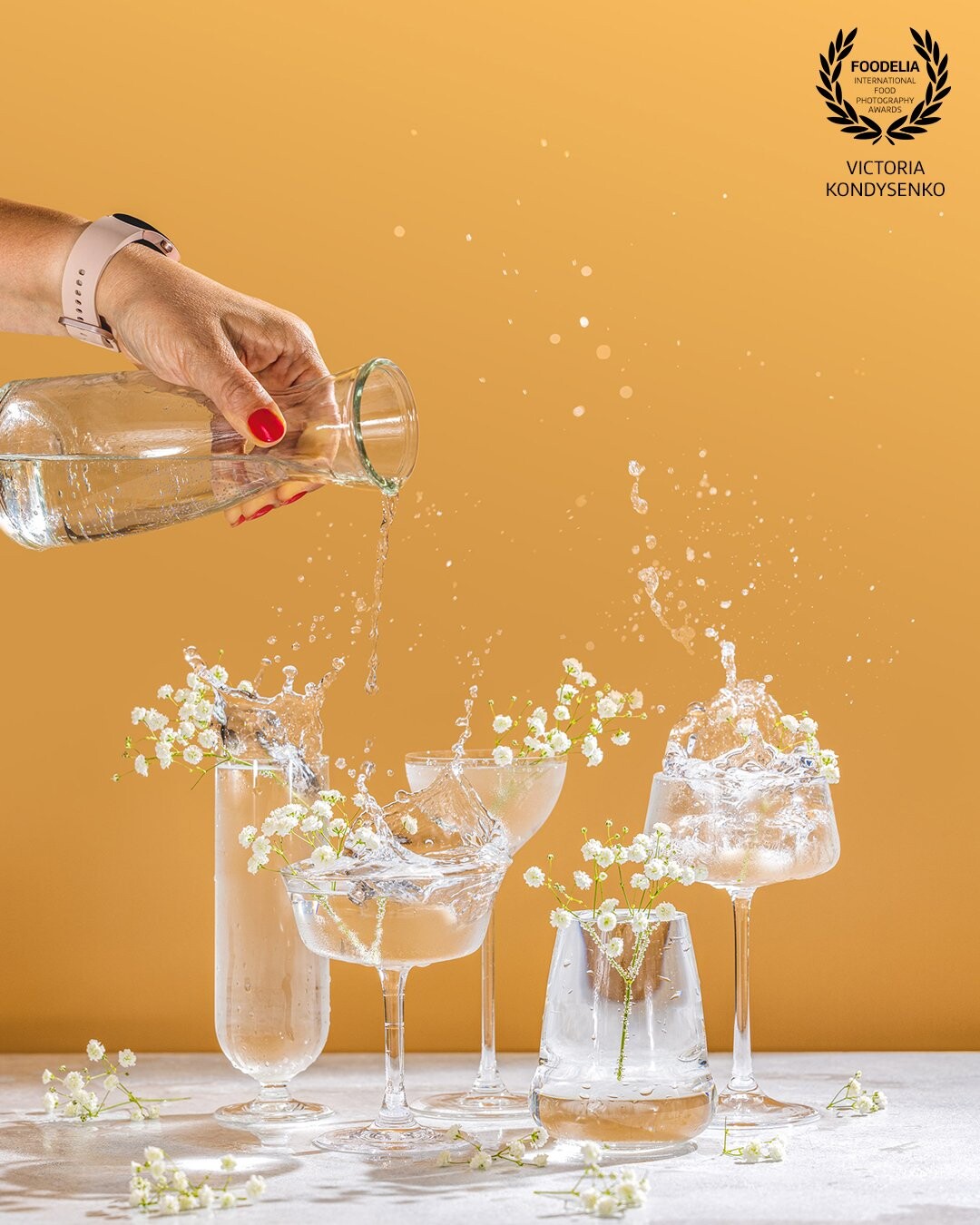 Various elegant champagne glasses with sparkling water, women hand pouring water from a bottle into a coupe glass. Splash, splash, fly flowers with water drops on peach color background