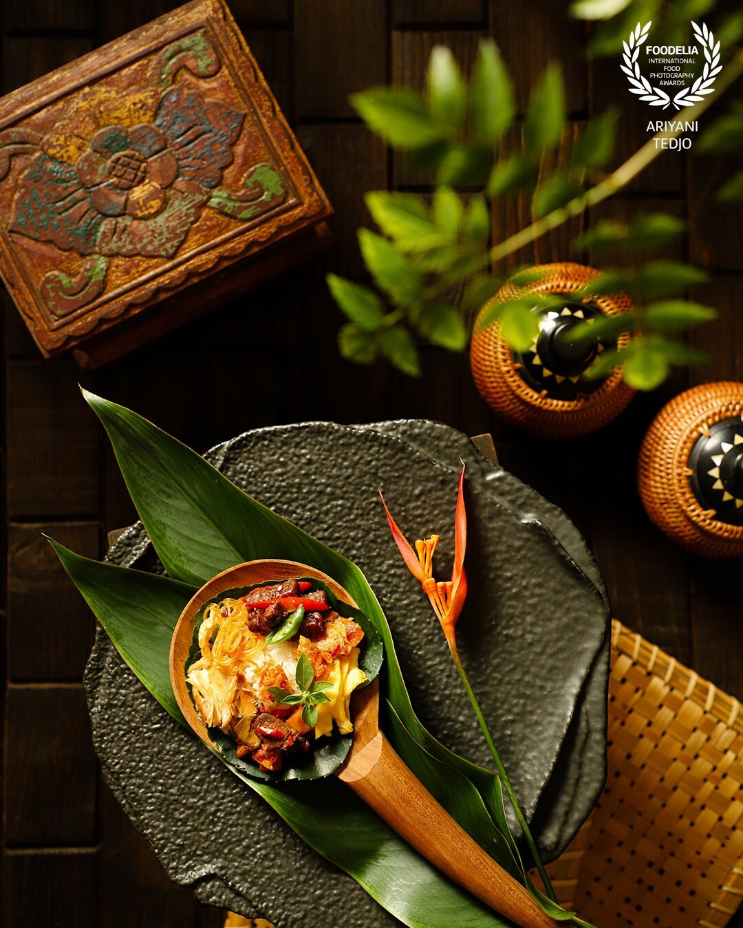 A traditional rice meal called Nasi Langgi, popular in Semarang, Central Java. Fragrant and savory rice topped with braised chicken, spicy liver stew, tempeh, glass noodles, potato crisps and omelet. This rice meal is served on a large wooden spoon lined with plum aralia leaf. Underneath the wooden spoon are two decorative banana leaves. The decorative banana blossom is placed on the plate next to the spoon.
