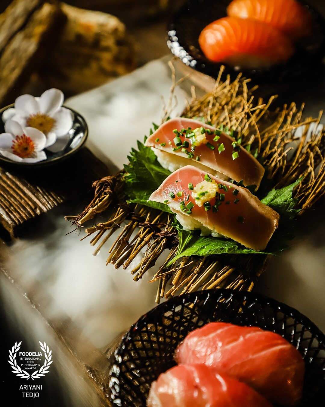 A nigiri sushi of lightly seared tuna. The sushi is placed on a mat of thin branches, lined with shiso leaves. The sushi is presented on wooden drawer set with smoke from dry ice.