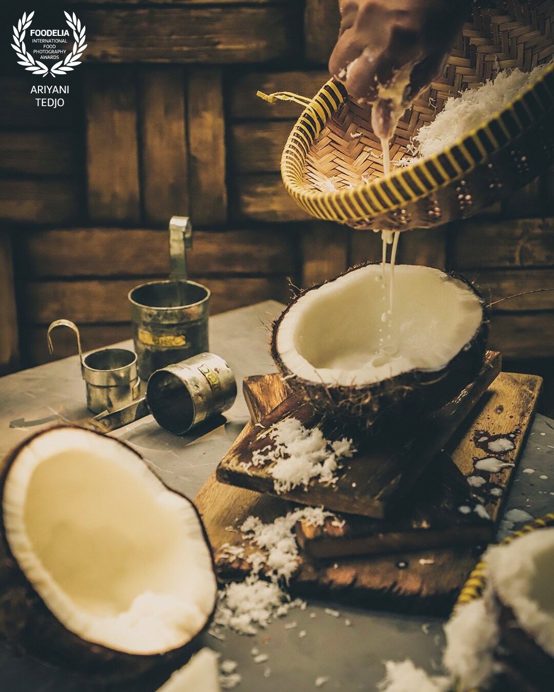Coconut milk is one signature ingredient in Indonesian cuisine. Using freshly made coconut milk is still preferred by some people, even though it is a pretty exhausting process. <br />
<br />
Here the milk is extracted by hand-squeezing the grated coconut that has been mixed with warm water. A traditional bamboo strainer specially made for coconut milk is used to catch the loose coconut from falling into the extracted liquid. A set of traditional tin measuring cups commonly used to measure coconut milk is shown in the scene.