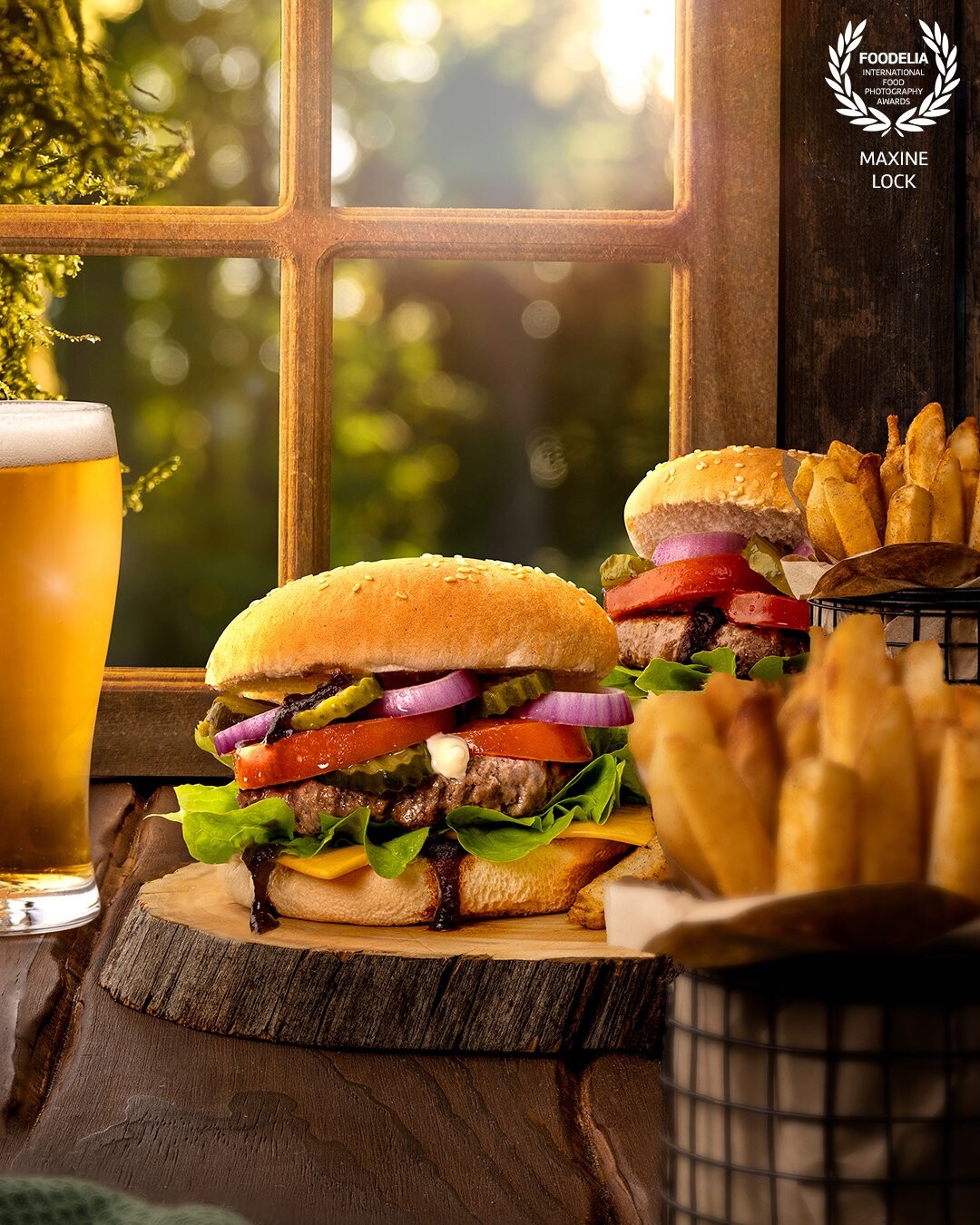 Burgers & fries, with a beer on the side, sitting on a table next to a window with sunlight pouring through; shining through the glass of beer.