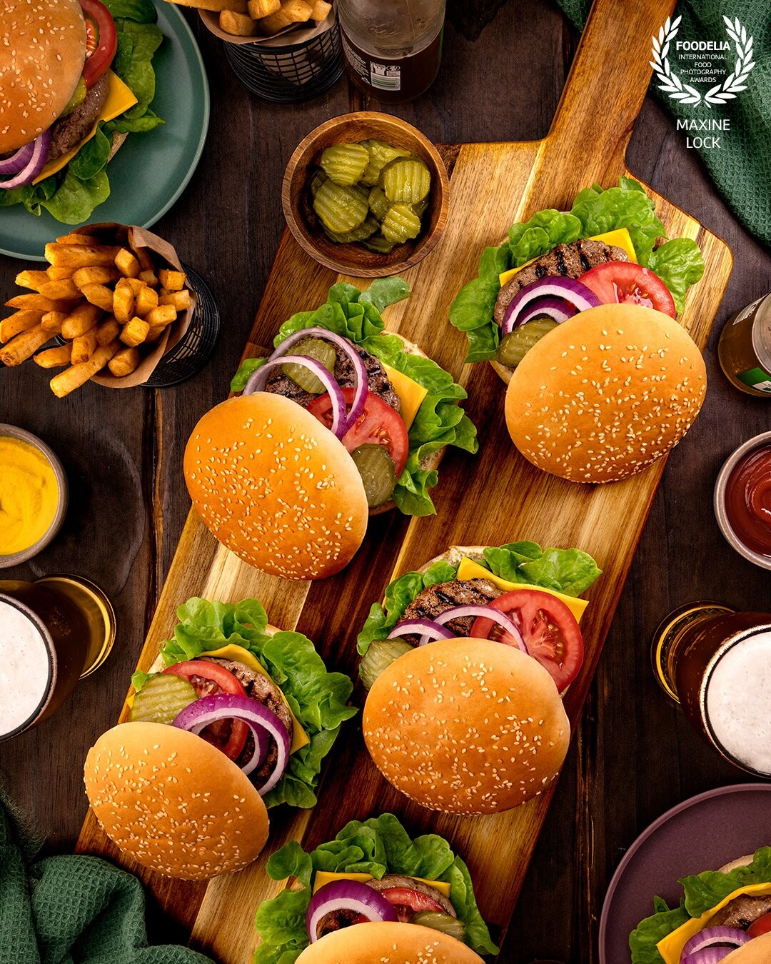 Multiple burgers on a wooden board, with the top bun slightly a skewed to show the inside fillings of the burgers. The table itself is surrounded by plates of burgers, fries, sauces and beer glasses and bottles.