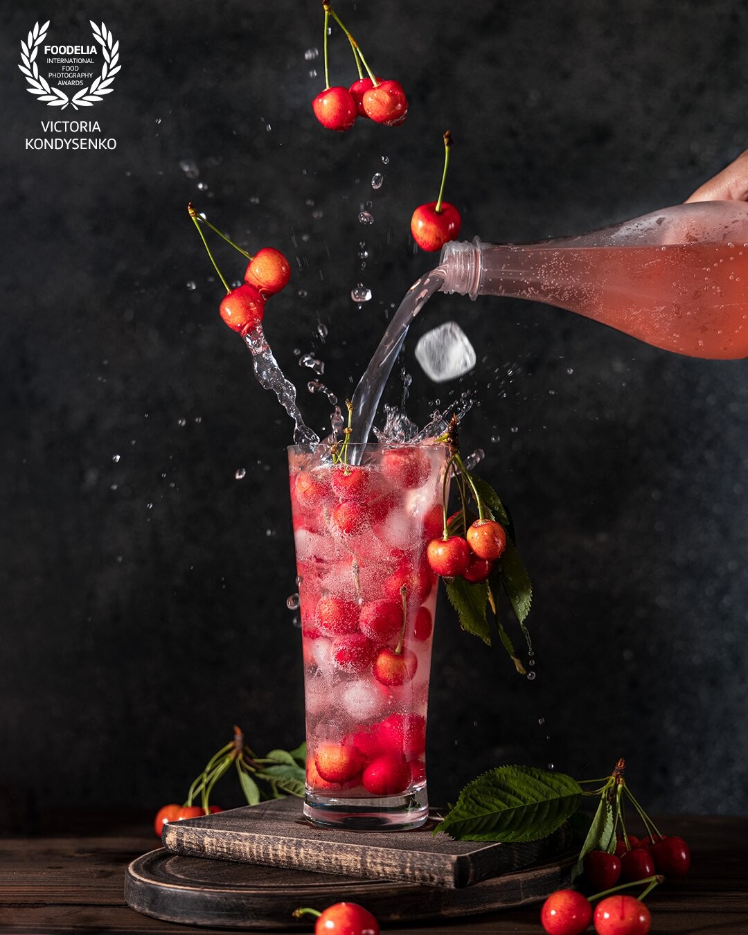 Glass of many cherry or merry berries with lemonade water splash, fly berries and ice, pouring lemonade from bottle to glass surround of fresh cherry or merry berries. Dark rustic wooden surface