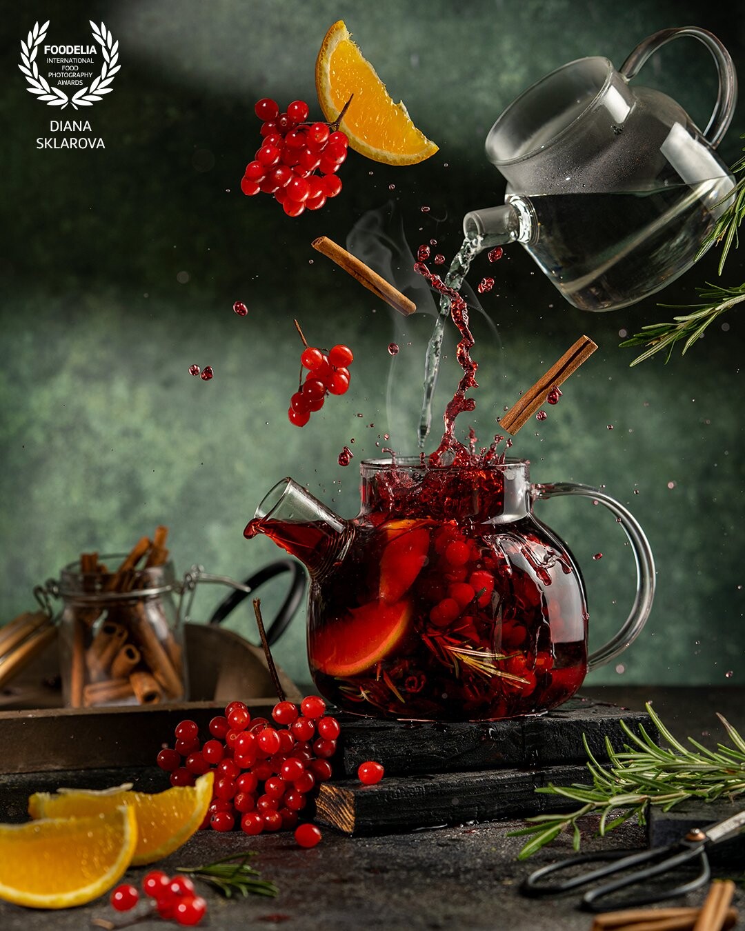 Spicy fruit tea with guelder rose, cinnamon, orange and herbs in transparent glass teapot on dark background. Levitation, flying  conceptual photo of healthy hot drink with splash and drops on dark green background.