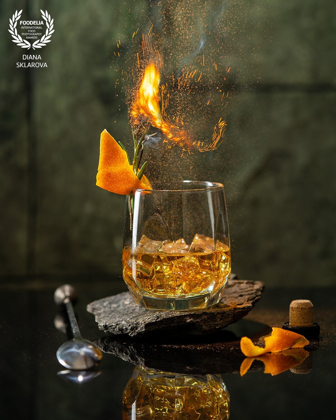 Whiskey in crystal glass with rosemary, ice cubes, orange and beautiful swirls of smoke. fire and burning cinnamon powder on dark green background.