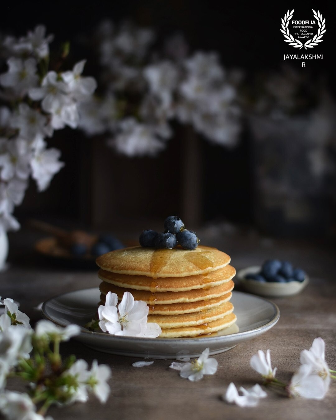 A regular breakfast scene . A stack of pancakes set amidst a floral setup . The idea was to capture the essence of spring by setting the scene with cherry blossom . Shot in natural light.