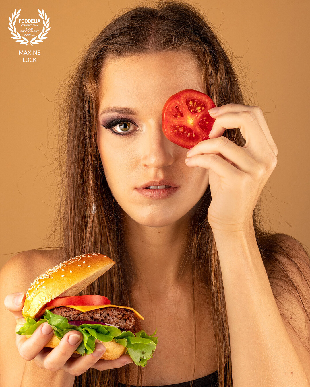 Model holding a burger with one hand, while the other hand is holding a tomato to her eye.