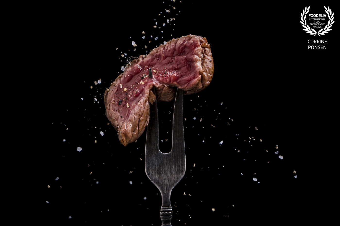 This beautyful juicy steak is grilled. A small piece of that steak is on top of a vintage fork. Himalaya salt is flying around the steak.