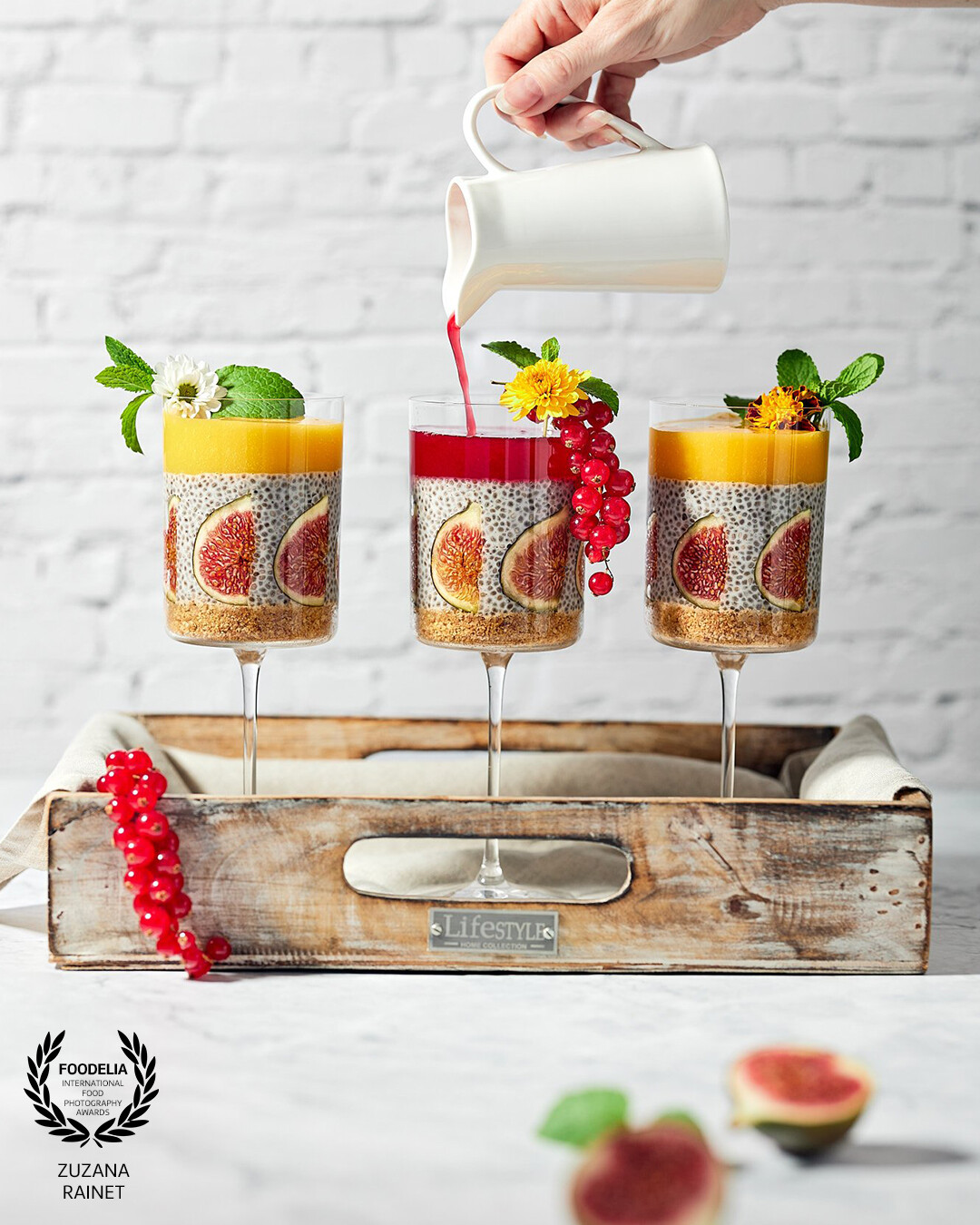 This image represents a modern eye catching styling of a coconut chia pudding with fresh figs and mango puree decorated with eatable flowers. <br />
<br />
I wanted the image to be minimalistic as the dessert itself is busy and colourful. The frozen movement of pouring liquid adds an interest to the whole frame while the light and airy style makes the dessert to stand out. Shot with studio flash light.