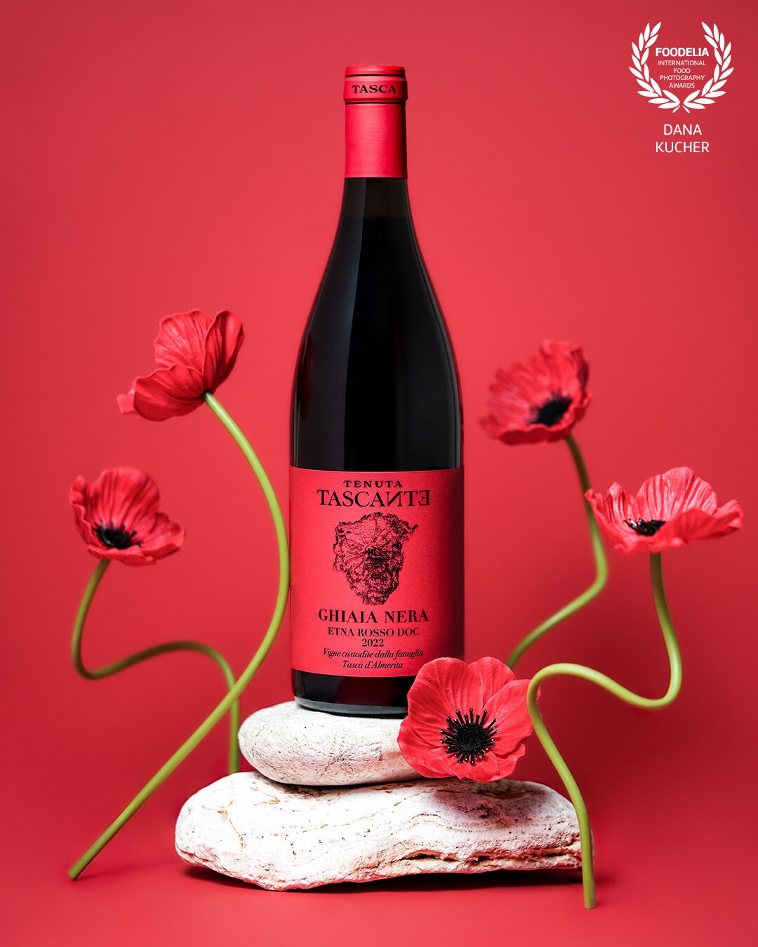 The Passion of Etna<br />
Deep as lava and elegant as poppies in the wind – this wine masterpiece, Ghiaia Nera Etna Rosso, embodies the energy and strength of Sicily. The stones beneath the bottle symbolize its volcanic origin, while the vibrant flowers evoke a sense of living passion. The rich red background enhances the bold character of the drink, as if inviting you to immerse yourself in a world of refined taste.