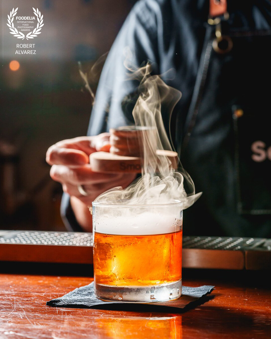 This photo captures a smoky Old Fashioned in the middle of its dramatic reveal at Loja Speakeast in Mayaguez, Puerto Rico. Shot with a strong key light from the side, the image freezes the swirling motion of the smoke as it lifts from the glass, highlighting the rich amber tones of the cocktail beneath.<br />
<br />
The texture of the smoke, combined with the clarity of the glass and the warm reflection off the bar, creates a moody, cinematic moment that feels both intimate and bold. The bartender’s hands in the background give it that behind-the-scenes energy—like you’re getting a front-row seat to the magic.