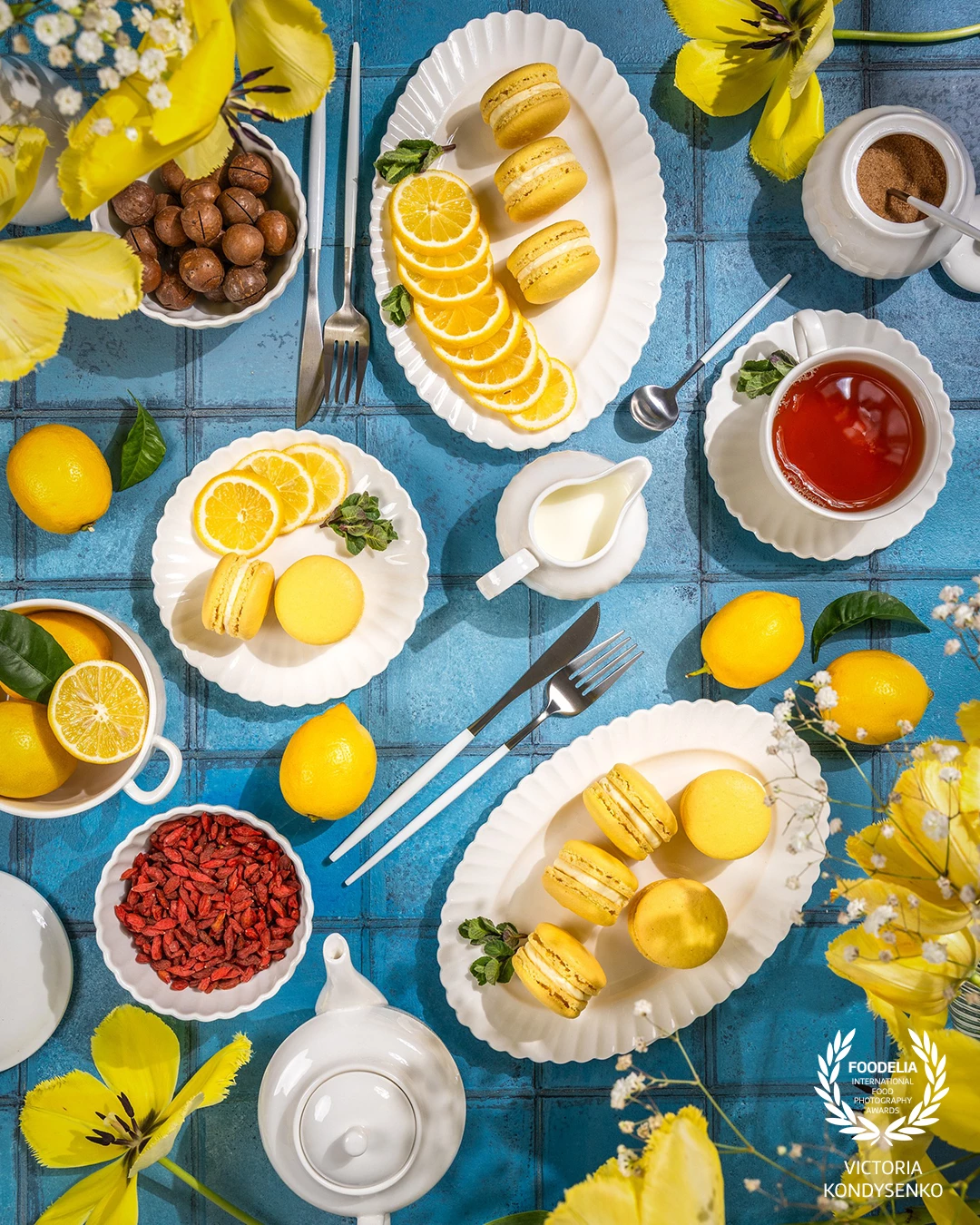 Delightful arrangement of lemon macarons, tea, and fresh lemons, accompanied by a bouquet of yellow tulips, sets a cheerful scene on a blue table.
