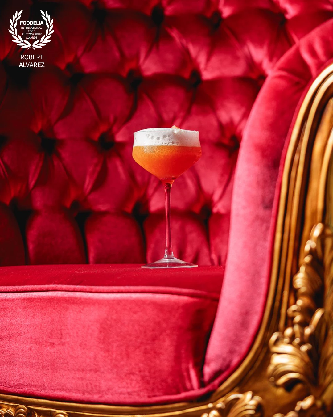 This photo, taken at Lojas Speakeasy in Mayagüez, Puerto Rico, captures the elegance and bold character of the space in a single frame. A vibrant cocktail sits perfectly centered on a luxurious red velvet armchair, its foam topping contrasting beautifully against the deep, tufted background and ornate golden frame.<br />
<br />
The rich textures and saturated colors bring a sense of drama and sophistication, while the clean composition lets the drink speak for itself. The shot feels regal, almost theatrical—highlighting not just the cocktail, but the essence of the speakeasy’s opulent and mysterious vibe.