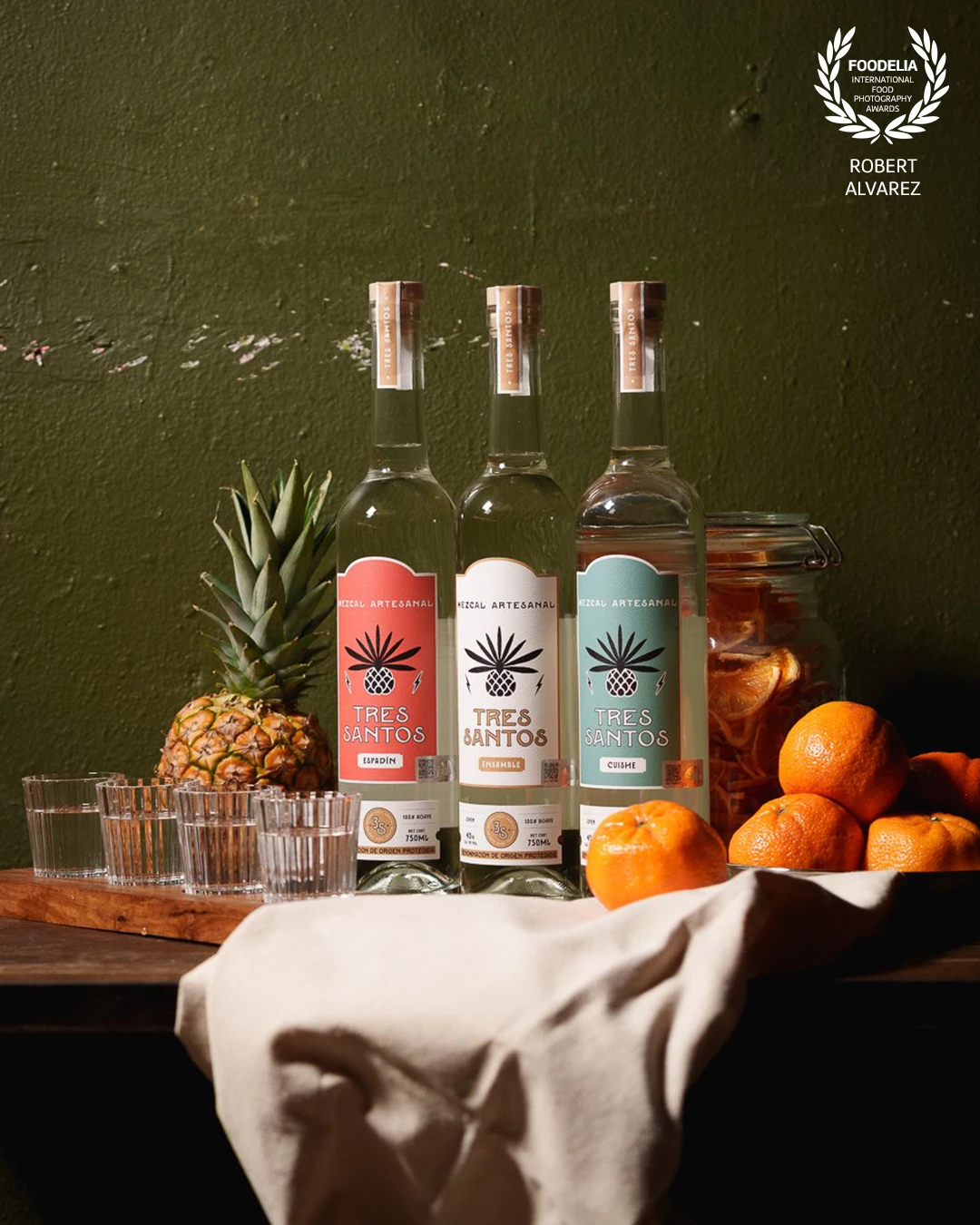 This photo, taken at Bar 0.2 in San Juan, Puerto Rico for Tres Santos Mezcal, was styled to feel warm, artisanal, and rooted in natural textures. For the lighting, I used a single directional softbox placed high and to the left, angled down to create strong highlights on the bottles and citrus while casting soft shadows to the right. This setup enhances the glass reflections and gives depth to the product labels without overwhelming them.<br />
<br />
A bounce card was used on the opposite side to softly lift the shadows and maintain detail, especially around the tangerines and cloth. The backdrop—a weathered olive green wall—adds texture and contrast while reinforcing the rustic, handcrafted vibe of the brand. Every element, from the wood and linen to the fruits and glassware, was chosen to reflect the earthy and bold identity of the mezcal itself.