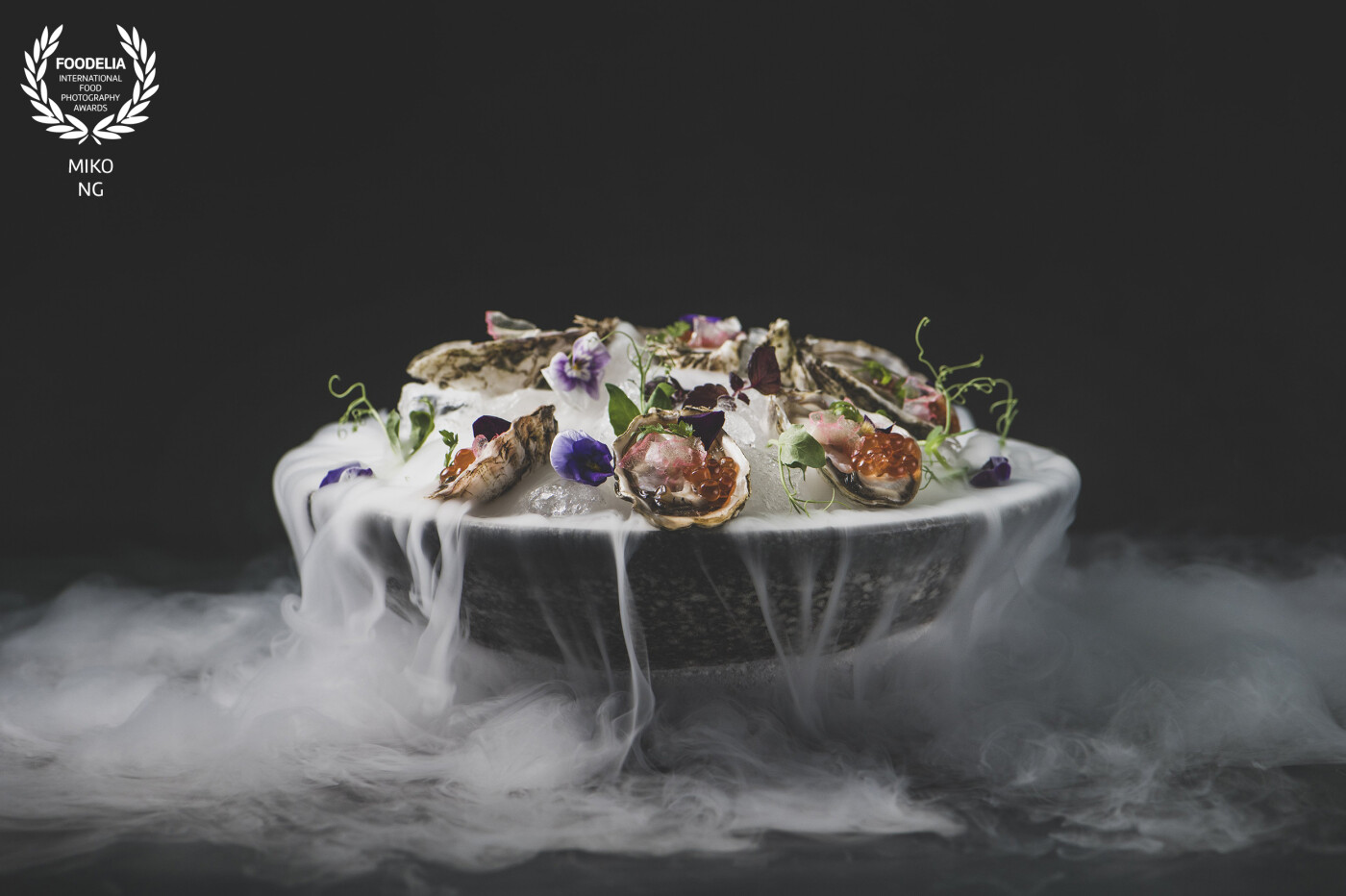A dark, elegant bowl presents fresh oysters nestled on a bed of crushed ice, adorned with edible flowers and delicate greens. Wisps of vapor rise gently, adding a dramatic, ethereal touch. Each oyster glistens with vibrant orange roe and colorful toppings, making the dish as visually captivating as it is refined.