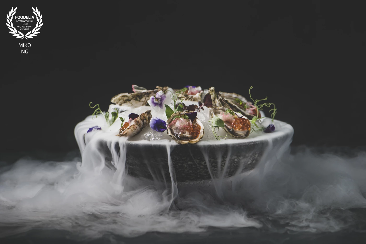 A dark, elegant bowl presents fresh oysters nestled on a bed of crushed ice, adorned with edible flowers and delicate greens. Wisps of vapor rise gently, adding a dramatic, ethereal touch. Each oyster glistens with vibrant orange roe and colorful toppings, making the dish as visually captivating as it is refined.