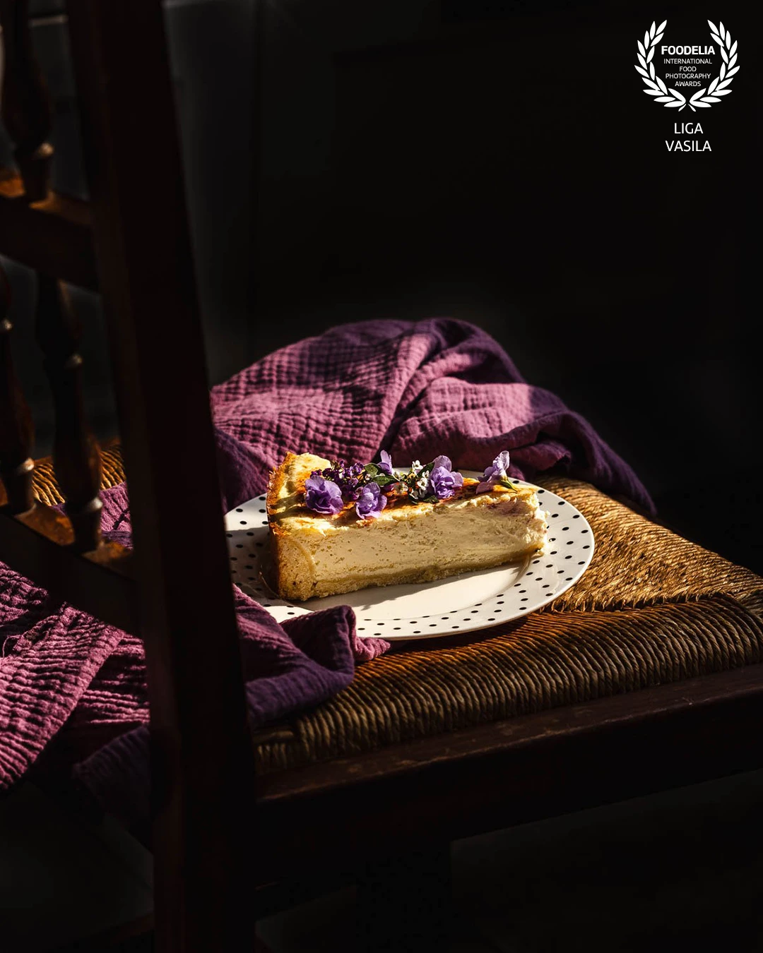 A slice of a home-baked cheesecake with a seemingly unintentional serving on a rustic chair. A sweet treat for someone special on a dreamy summer evening, framed with the directional lines of the chair seat and the wavy fabric of a matching color.