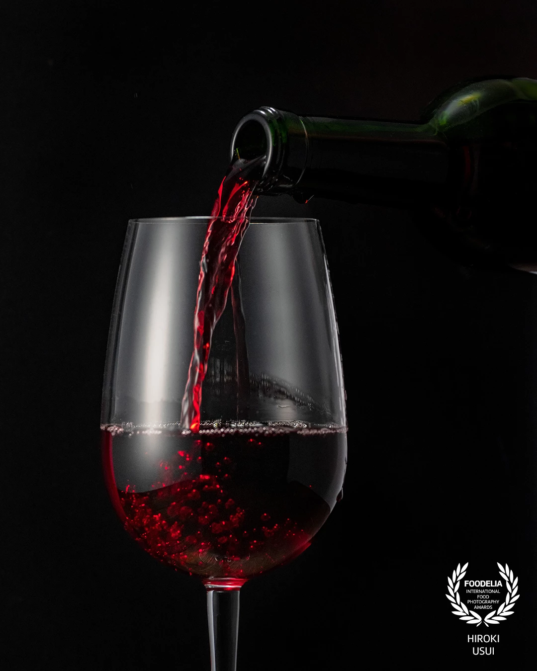 This photograph freezes the elegant moment of red wine being poured into a glass. The black background creates a strong contrast, making the rich red tones of the wine stand out. Carefully controlled lighting highlights the texture of the liquid, the clarity of the glass, and the dynamic motion of the pour.<br />
<br />
Bubbles rising inside the wine add a sense of freshness and movement, while the composition keeps the focus simple and refined. A clean, minimal setup with high-speed capture turns an everyday moment into a visually striking image