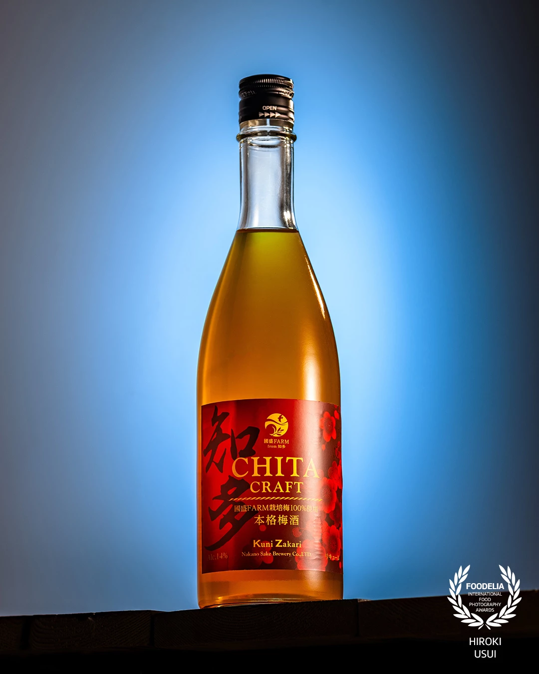 This image of Chita Craft Umeshu showcases the artistry of product photography through meticulous lighting and composition. A smooth gradient blue background is used as a backlight, allowing the amber color of the plum liqueur to glow with clarity and warmth. A subtle top-side light highlights the curvature of the glass bottle, adding dimension and transparency.<br />
<br />
The bold red and gold label is carefully illuminated to enhance its vibrancy without overpowering the overall balance. Light falloff and shadow control are precisely managed to retain the richness of the bottleโs texture while ensuring the label remains crisp and legible.<br />
<br />
Positioned on a dark base, the bottle casts a soft, grounded shadow that adds depth and visual stability. This composition masterfully balances color contrast, material expression, and spatial tension, making it a standout example of how photography can convey both product quality and brand story in a single frame.