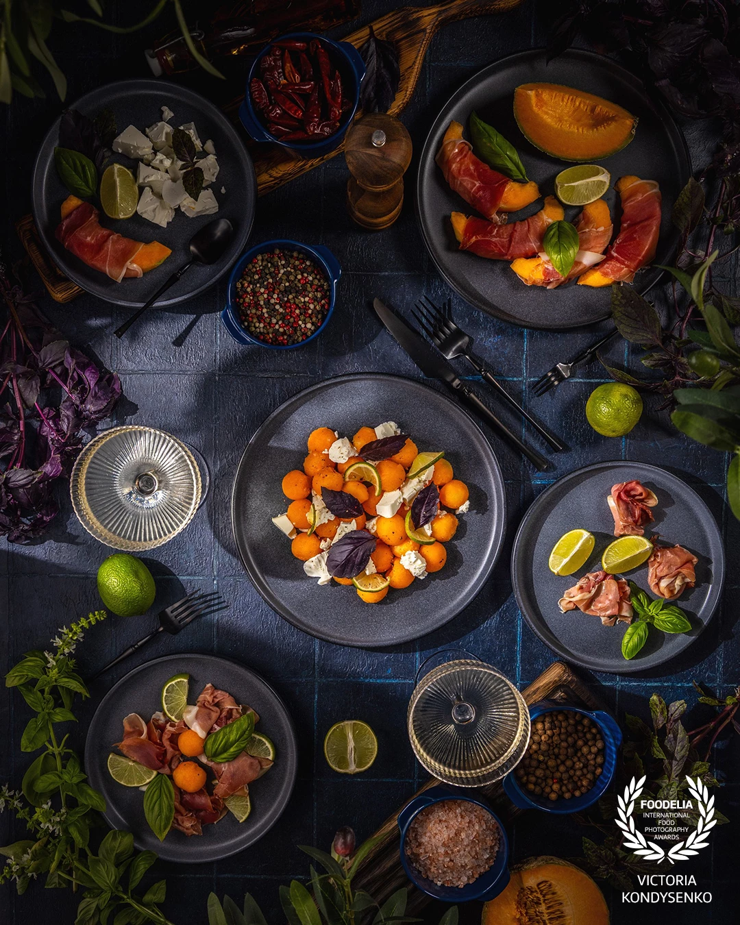 Plates of melon and prosciutto appetizer with feta cheese, basil, lime, and spices, arranged on a dark blue textured surface, creating a vibrant culinary scene
