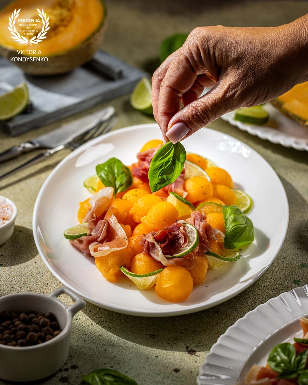 Chef is adding a fresh basil leaf to a vibrant melon salad, featuring cantaloupe balls, prosciutto, and lime, creating a colorful and appetizing dish