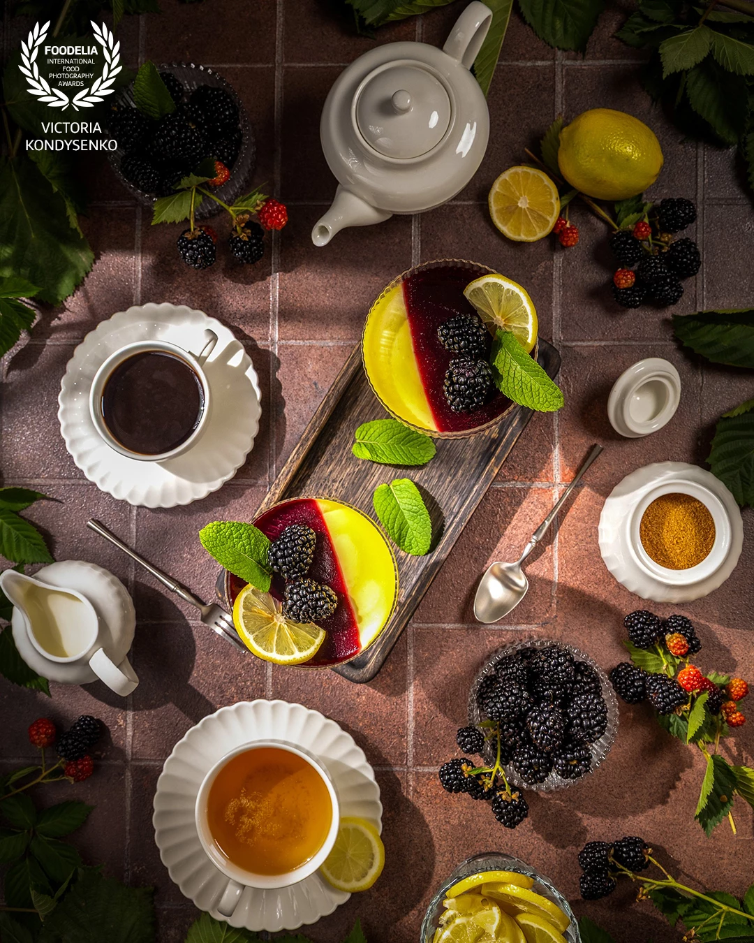 Two layered desserts with blackberries, lemon slices, and mint leaves are served on a rustic table alongside cups of tea and coffee, creating a delightful teatime scene