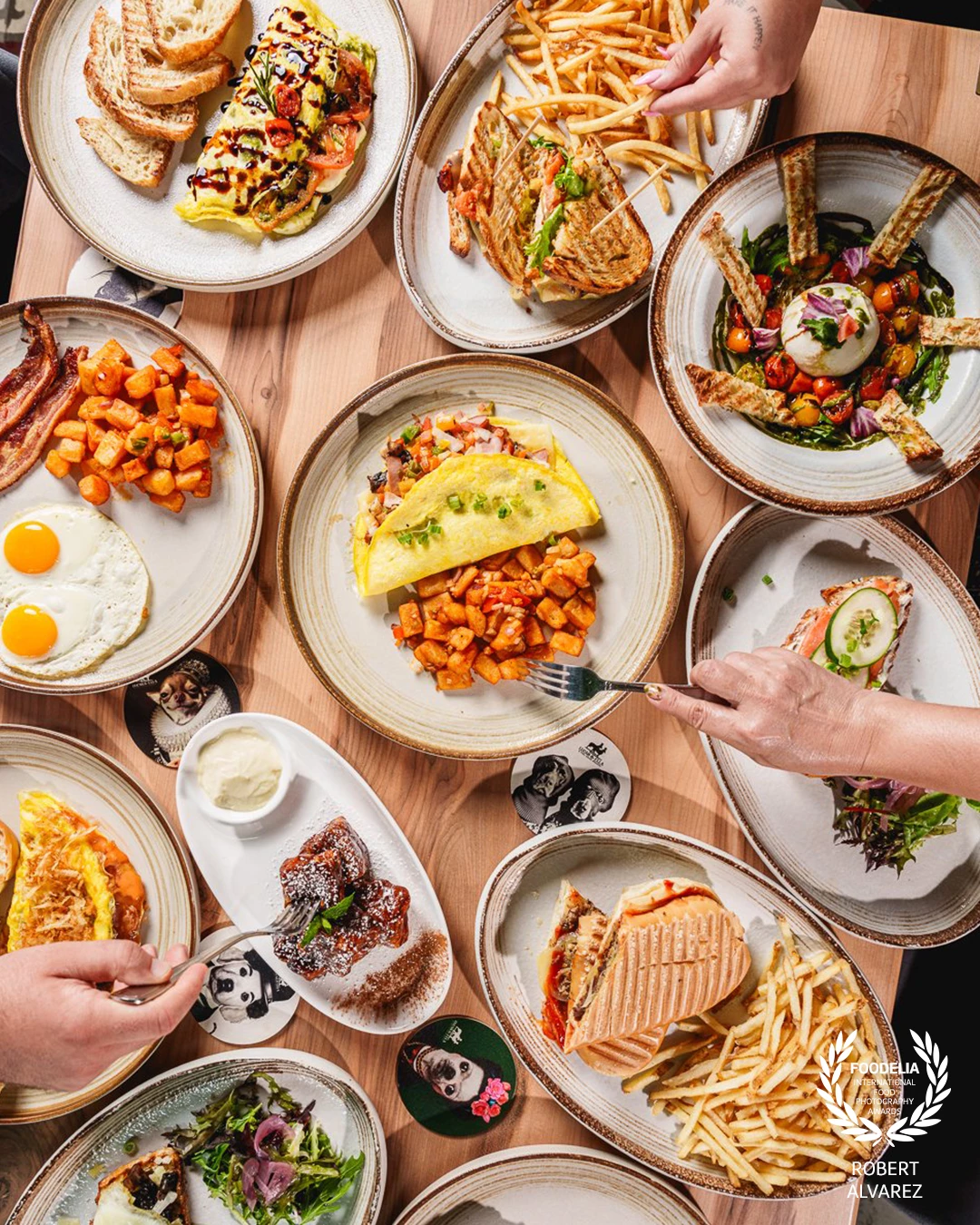 This vibrant overhead shot captures the brunch spread at Lucas & Ella in Aguadilla, Puerto Rico, where every plate tells its own story. From fluffy omelets paired with golden potatoes, to crispy paninis stacked with fries, to fresh burrata with colorful tomatoes and toasted bread, the table bursts with variety and texture. The inclusion of hands reaching for food adds life and movement, inviting the viewer into the scene as if they were about to join the meal.<br />
<br />
The composition emphasizes abundance and togetherness—perfectly reflecting the joy of brunch shared among friends. Warm light and careful framing bring harmony to the organized chaos of the table, making the food look not only delicious but also inviting.