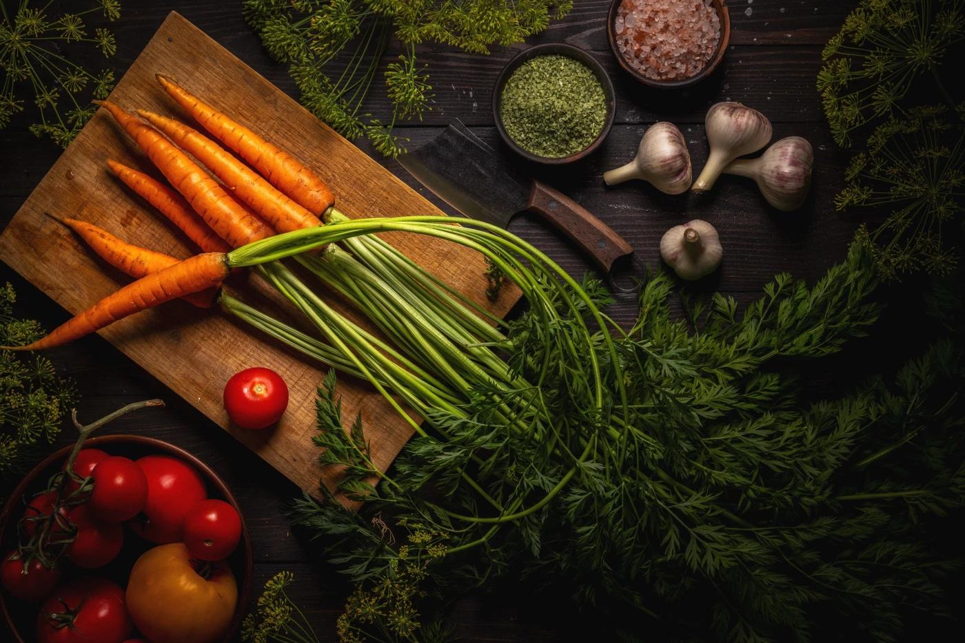 Rustic wooden board displaying a bunch of fresh orange carrots with vibrant green tops. Ripe red tom...