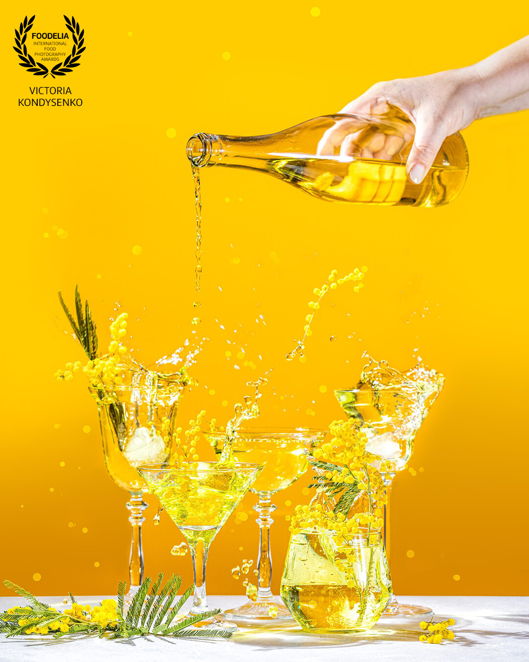 Various elegant martini and whiskey glasses with sparkling water, womans hand pouring water from a bottle into a coupe glass. Splash, splash, fly yellow mimosa flowers with water drops on yellow background color.