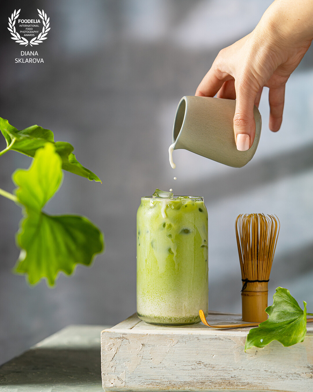 Pouring milk in matcha cold tea drink, grey background with fresh leaves and beautiful  sunny light. Healthy vegan drink.